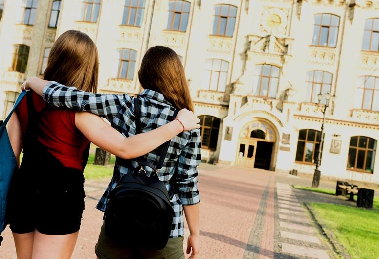 Two girls with backpacks hugging in front of a high school