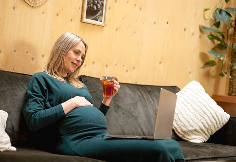 A pregnant woman in a green dress drinks tea on a sofa